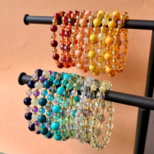 May include: A display of beaded bracelets in various colors, including red, orange, yellow, blue, and green. The bracelets are strung on elastic and feature a variety of bead sizes and materials. The bracelets are displayed on a black metal stand.