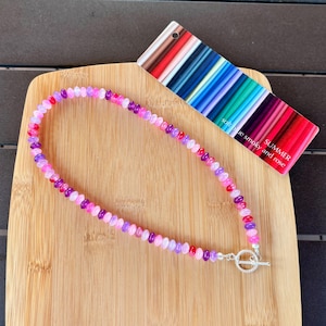 May include: A necklace made of small, round, translucent beads in shades of pink, purple, and magenta. The necklace features a silver-toned toggle clasp and is displayed next to a rectangular color swatch card labeled SUMMER.