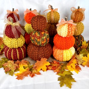 May include: A collection of stacked, handcrafted pumpkins in various colors, including orange, yellow, and burgundy. Each pumpkin is made of knitted yarn and topped with a small twig and ribbon. Autumn leaves surround the pumpkins.