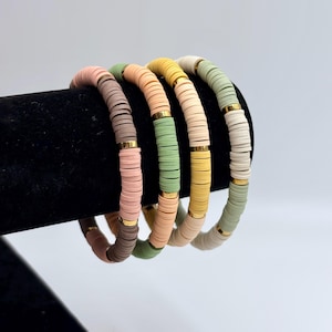 May include: Four colorful beaded bracelets displayed on a black surface. The bracelets feature flat, round beads in shades of pink, brown, green, yellow, and cream, separated by gold-colored metal spacers. The bracelets are a stylish accessory.