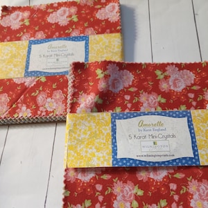 May include: A stack of red fabric squares with a white floral pattern. The fabric squares are labeled "Amorette by Kaye England 5 Karat Mini-Crystals Wilmington www.wilmingtonprints.com"