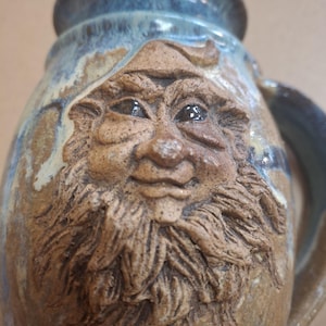 May include: A handcrafted ceramic mug with a detailed, sculpted face of a bearded man. The mug features a brown and tan color scheme with blue accents around the rim and handle. The face has a whimsical, folk art style.