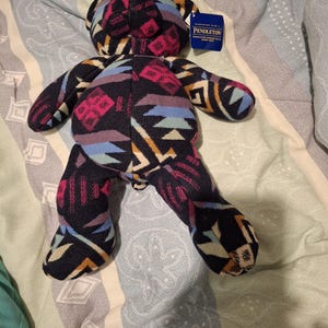 Può includere: Un orsacchiotto di peluche Pendleton con un colorato motivo geometrico nei toni del nero, rosa, blu e giallo. L'orso ha un'etichetta con il logo Pendleton. L'orso è sdraiato su un copriletto fantasia.