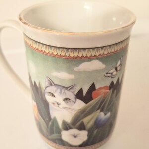 May include: A white ceramic mug featuring a decorative band at the top and bottom with a gold-toned rim. The central illustration depicts a white and grey cat nestled among green foliage, colorful flowers, and a small butterfly under a pale sky.