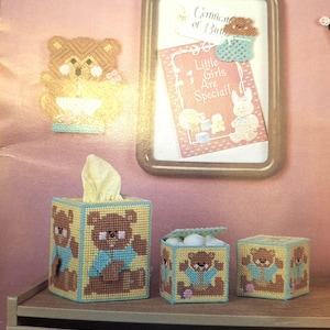 May include: A set of three plastic canvas items featuring brown teddy bears wearing blue shirts on a yellow background. The set includes a tall rectangular tissue box cover, a small square lidded box, and a small square box, all with matching bear motifs.