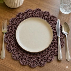May include: A round, dark brown crocheted placemat with a scalloped edge, featuring a central beige ceramic plate. A silver-toned metal fork is positioned to the left, while a silver-toned metal knife and spoon are placed to the right of the plate.