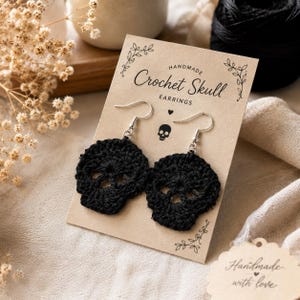 May include: A pair of black crochet skull earrings attached to a tan card with the text Handmade Crochet Skull EARRINGS and a small skull icon. The earrings feature silver-toned metal hooks and are crafted from black thread in a skull shape.