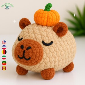 May include: A crocheted capybara plush toy with a small orange pumpkin on its head. The capybara is tan with brown accents on its ears, face, and feet. The pumpkin has a green stem. The toy has a smiling face.