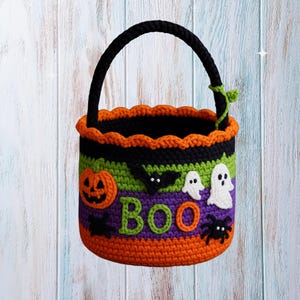 May include: A handmade crochet Halloween basket in orange, black, green, and purple. The basket features a pumpkin, ghosts, bats, and a spider, with the word "BOO" in green letters. It has a black handle and orange scalloped trim.
