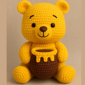 May include: A handmade, yellow crochet bear with a brown honey pot. The bear has black eyes and a smiling face. The honey pot is brown with a yellow honey dripping design. The bear is sitting.