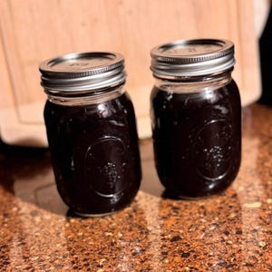 Könnte beinhalten: Zwei Gläser mit einer dunklen, zähflüssigen Flüssigkeit, möglicherweise Konserven oder eine Sauce. Die Gläser haben silberne Metalldeckel und stehen auf einer gesprenkelten, braunen Oberfläche. Die Gläser stehen eng beieinander.