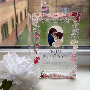Puede incluir: Decoración de libro de acrílico transparente con el título "Pride & Prejudice" y una ilustración de una pareja. El diseño incluye detalles florales y una cita de Jane Austen. El logotipo de De Ember es visible.