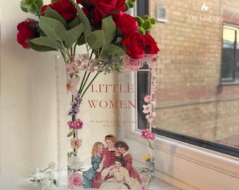 May include: A clear acrylic vase featuring the book cover of "Little Women" with a floral border. The vase holds red roses and greenery, creating a decorative display. The vase is placed on a windowsill.