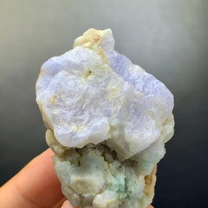 May include: A natural mineral specimen featuring a cluster of light purple and off-white crystalline formations. The rock has a rough, textured surface with hints of green and brown.