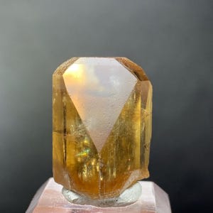 May include: A translucent, golden-yellow faceted gemstone crystal with a hexagonal prismatic shape. The stone features a flat, polished top facet and vertical side faces, displaying internal clarity and light reflection throughout its structure.