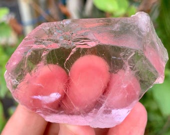 May include: A translucent, rough-cut, pink crystal with a faceted surface. The crystal has a soft, rosy hue and appears to be held in a hand, with a blurred green background. The stone has a natural, organic shape and texture.