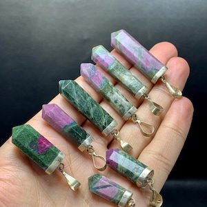 May include: A collection of gemstone pendants with silver-colored metal findings. The gemstones are a mix of green and pink hues, with a faceted, rectangular shape. The pendants are held in a hand against a dark background.
