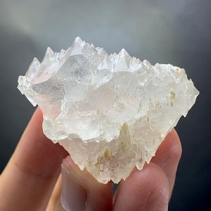 Può includere: Un gruppo di cristalli di fluorite trasparente con formazioni appuntite e affilate. Il minerale ha una qualità traslucida, con sottili variazioni di colore e consistenza. La forma generale è irregolare, con un aspetto ruvido e naturale. La fluorite è tenuta in una mano.