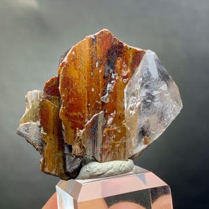 Puede incluir: Un espécimen mineral natural con cristales marrones, naranjas y transparentes. Los cristales están montados sobre un soporte de exhibición rectangular transparente. El mineral tiene una apariencia en capas con un cristal de cuarzo translúcido y vítreo.