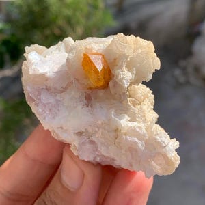 Puede incluir: Primer plano de un espécimen mineral en bruto y natural. El espécimen presenta una vibrante formación cristalina de color naranja-amarillo, incrustada en una matriz de minerales blanquecinos y rosa pálido. La textura rugosa y las variaciones de color resaltan su origen natural.