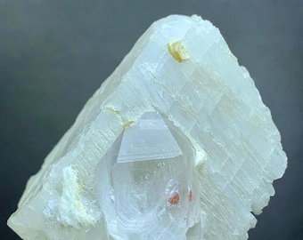 May include: A light blue crystal formation with a clear, pointed crystal embedded in the center. The crystal has a rough, textured surface with a few small, yellow-brown spots.