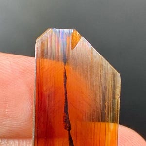 Puede incluir: Una gema rectangular y translúcida con una paleta de colores cálidos. La piedra presenta una mezcla de tonos naranja, rojo y marrón, con una inclusión lineal oscura que atraviesa su centro. La superficie tiene una textura estriada y los bordes están facetados.