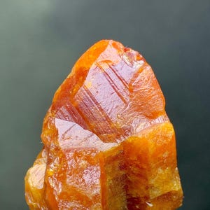 May include: A vibrant orange mineral specimen with a crystalline structure. The rough-hewn stone is mounted on a clear, rectangular base. The mineral's surface reflects light, showcasing its natural texture and color variations.