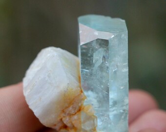 May include: A light blue aquamarine crystal with a rectangular shape, next to a white and orange crystal. The aquamarine has a clear, glassy appearance. The crystals are held in a hand.