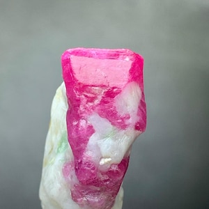Pode incluir: Um cristal de rubi rosa com inclusões brancas. O cristal é bruto e tem um aspecto natural não polido. Está montado numa base cinzenta.