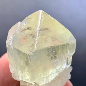 Könnte beinhalten: Ein durchscheinender, hellgelber Kristall mit pyramidenförmiger Spitze und rauer, kristalliner Basis. Die Facetten des Kristalls fangen das Licht ein und zeigen seine natürliche Struktur. Die Gesamtform ist geometrisch, mit einer leicht rauen Textur.