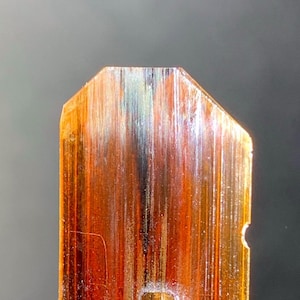 Puede incluir: Una gema rectangular translúcida con una superficie pulida. La piedra muestra un degradado de colores, incluyendo ámbar, marrón y toques de verde y negro. Está montada sobre un soporte de metal plateado.