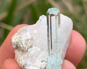 May include: A natural aquamarine crystal with a light blue hue, set against a white quartz matrix. The crystal is a prismatic shape with vertical striations. The base of the crystal is attached to a cluster of silvery-gray mica.