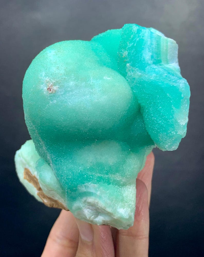 Puede incluir: Un vibrante esp&eacute;cimen mineral de color turquesa, con una forma redondeada y burbujeante. La superficie tiene un aspecto texturizado, con &aacute;reas de tonos turquesa m&aacute;s claros y oscuros. La base del mineral tiene un borde &aacute;spero y marr&oacute;n.