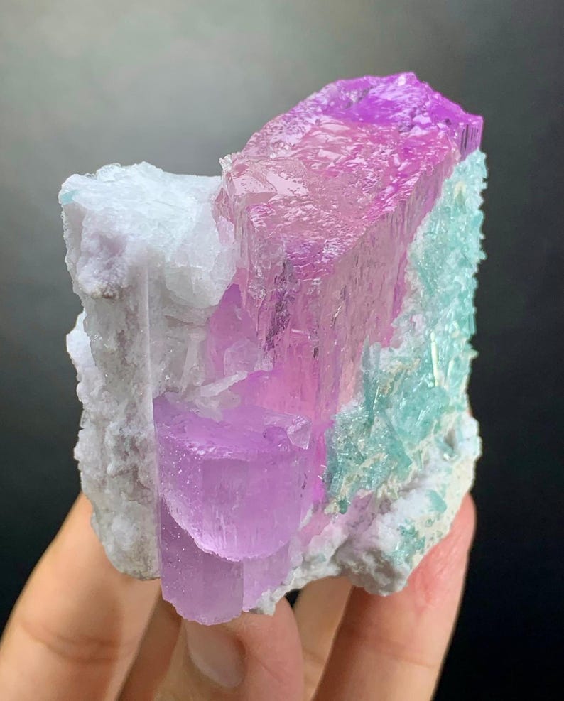 K&ouml;nnte beinhalten: Ein rohes Mineralexemplar mit durchscheinenden, leuchtend rosa und violetten Kristallstrukturen, die sich von einer texturierten, undurchsichtigen wei&szlig;en Matrix abheben. Die Gruppe zeigt zudem Bereiche mit hellt&uuml;rkisen Kristallformationen.