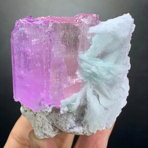 K&ouml;nnte beinhalten: Ein rohes Mineralexemplar mit einem gro&szlig;en, durchscheinenden, rechteckigen rosa Kristallabschnitt, der an einem B&uuml;schel blass minzgr&uuml;ner, klingenartiger Formationen befestigt ist. Die Basis des Exemplars ist eine raue, hellgraue, texturierte Gesteinsmatrix.