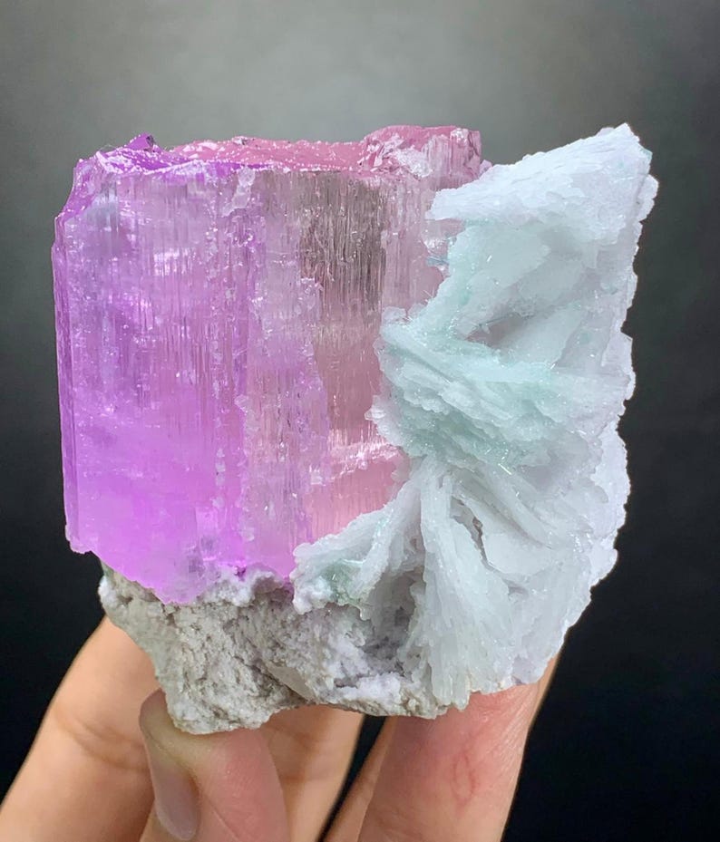 K&ouml;nnte beinhalten: Ein rohes Mineralexemplar mit einem gro&szlig;en, durchscheinenden, rechteckigen rosa Kristallabschnitt, der an einem B&uuml;schel blass minzgr&uuml;ner, klingenartiger Formationen befestigt ist. Die Basis des Exemplars ist eine raue, hellgraue, texturierte Gesteinsmatrix.
