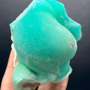 Puede incluir: Un vibrante esp&eacute;cimen mineral de color turquesa con una superficie redondeada y texturizada. La estructura cristalina es visible. El mineral se sostiene en una mano sobre un fondo oscuro. La forma es irregular y org&aacute;nica.