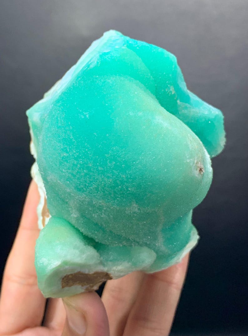 Puede incluir: Un vibrante esp&eacute;cimen mineral de color turquesa con una superficie redondeada y texturizada. La estructura cristalina es visible. El mineral se sostiene en una mano sobre un fondo oscuro. La forma es irregular y org&aacute;nica.