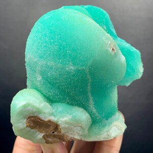 Puede incluir: Un vibrante esp&eacute;cimen mineral de color turquesa con una forma redondeada &uacute;nica. La superficie tiene una apariencia texturizada y cristalina, con &aacute;reas de color verde claro y marr&oacute;n. El mineral se sostiene en una mano, mostrando su belleza natural y caracter&iacute;sticas geol&oacute;gicas.