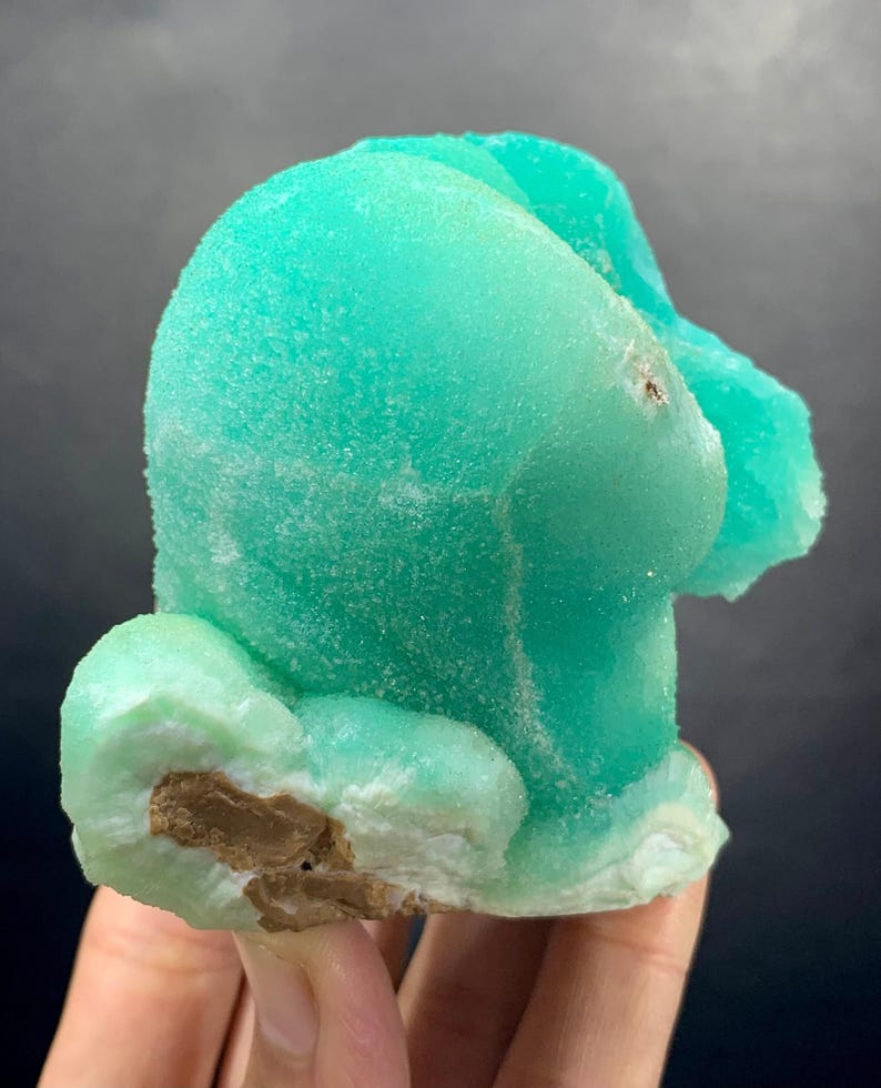 Puede incluir: Un vibrante esp&eacute;cimen mineral de color turquesa con una forma redondeada &uacute;nica. La superficie tiene una apariencia texturizada y cristalina, con &aacute;reas de color verde claro y marr&oacute;n. El mineral se sostiene en una mano, mostrando su belleza natural y caracter&iacute;sticas geol&oacute;gicas.