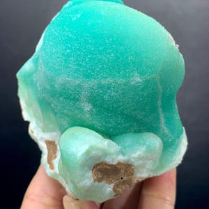 Puede incluir: Un esp&eacute;cimen mineral de color turquesa vibrante con una superficie texturizada. El mineral tiene una forma redondeada y org&aacute;nica con &aacute;reas de color marr&oacute;n claro y blanco en la base. El fondo es de color oscuro y s&oacute;lido.