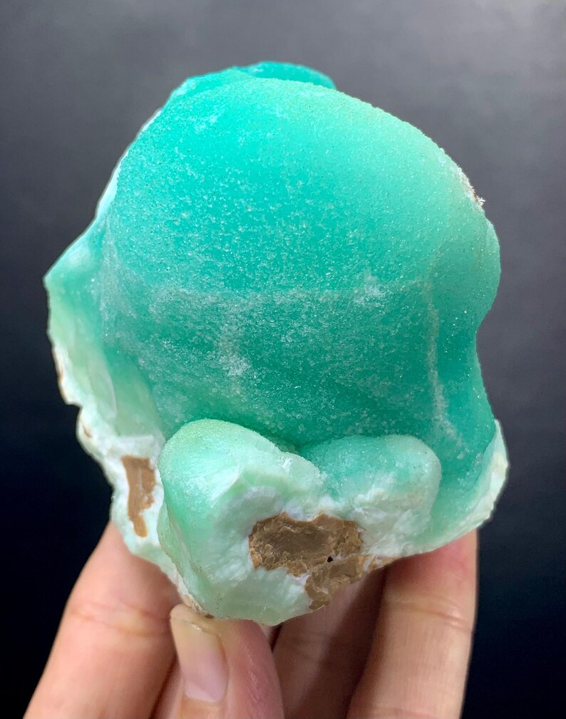 Puede incluir: Un esp&eacute;cimen mineral de color turquesa vibrante con una superficie texturizada. El mineral tiene una forma redondeada y org&aacute;nica con &aacute;reas de color marr&oacute;n claro y blanco en la base. El fondo es de color oscuro y s&oacute;lido.