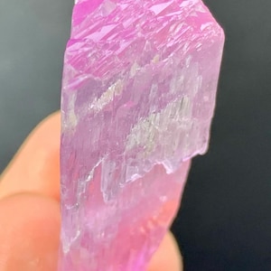 May include: A pink, translucent crystal with a striated surface. The crystal is pointed at one end and has a rough, uneven texture.