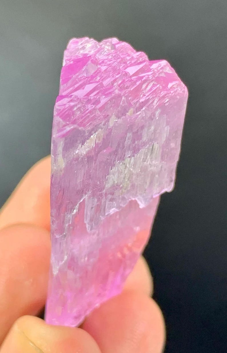 May include: A pink, translucent crystal with a striated surface. The crystal is pointed at one end and has a rough, uneven texture.