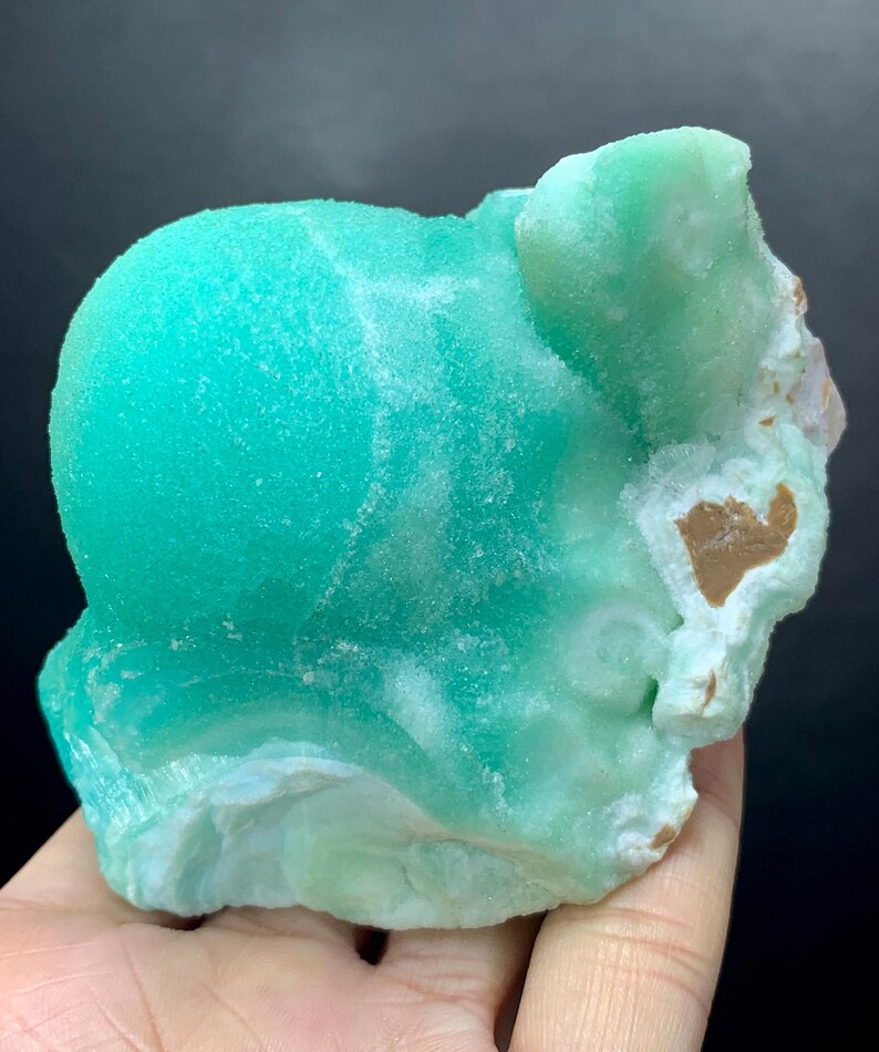 Puede incluir: Un esp&eacute;cimen mineral de color turquesa vibrante con una textura redondeada y burbujeante. La piedra tiene &aacute;reas blancas y marrones, y una apariencia rugosa y natural. El mineral se sostiene en una mano, mostrando su tama&ntilde;o y variaciones de color.