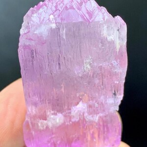 May include: A pink, translucent, faceted crystal with a pointed top and a flat base. The crystal has a striated texture and is approximately 2 inches tall.