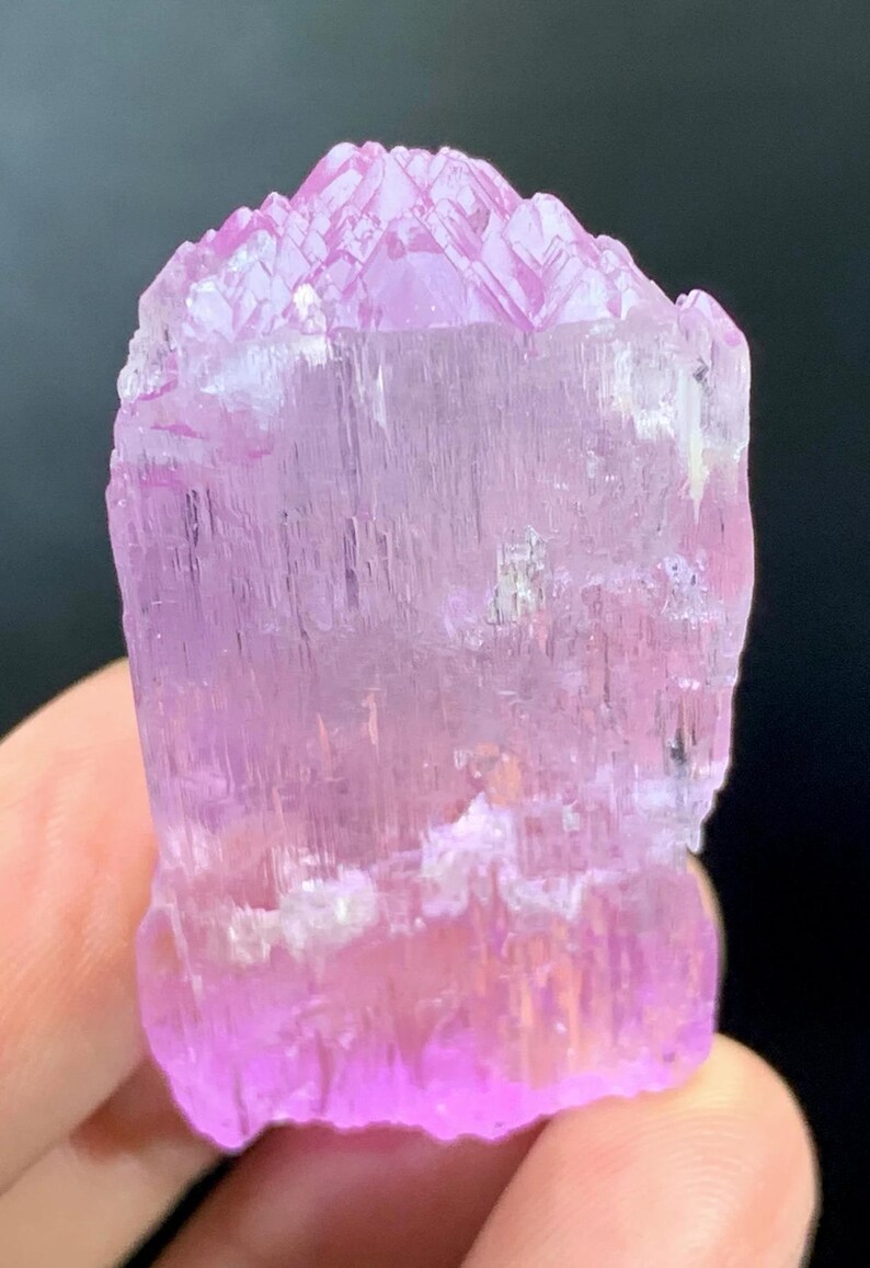 May include: A pink, translucent, faceted crystal with a pointed top and a flat base. The crystal has a striated texture and is approximately 2 inches tall.