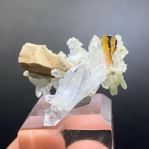 Puede incluir: Un grupo de cristales de cuarzo transparente con una formación rocosa marrón claro y un cristal naranja oscuro y negro. Los cristales están montados sobre una base rectangular rosa. El fondo es oscuro.
