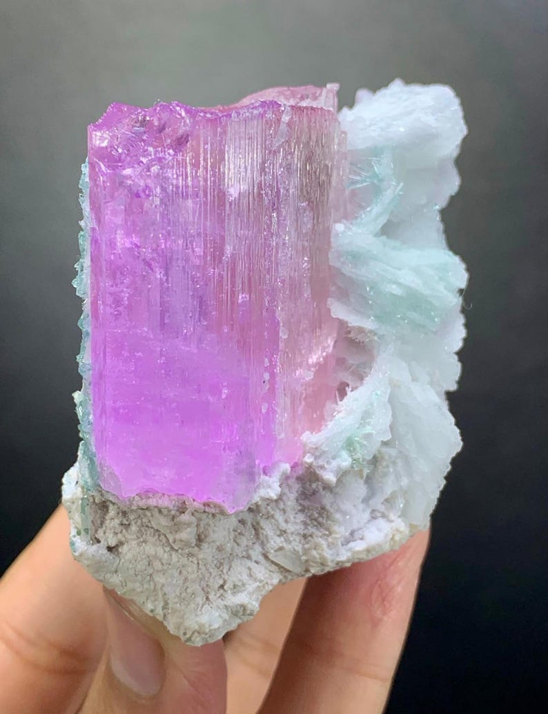K&ouml;nnte beinhalten: Ein rohes Mineralexemplar mit einer gro&szlig;en, durchscheinenden, vertikal gestreiften rosa Kristalls&auml;ule, die an einer Ansammlung wei&szlig;er und blass minzgr&uuml;ner Kristallformationen auf einer rauen, hellen Gesteinsbasis befestigt ist.