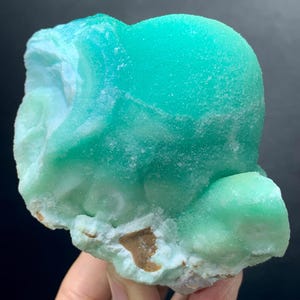 Puede incluir: Un vibrante esp&eacute;cimen mineral cristalino con un llamativo tono turquesa. El mineral tiene una forma redondeada y bulbosa con una superficie texturizada, y una base de color m&aacute;s claro, blanquecino y marr&oacute;n. La apariencia general es suave y pulida.